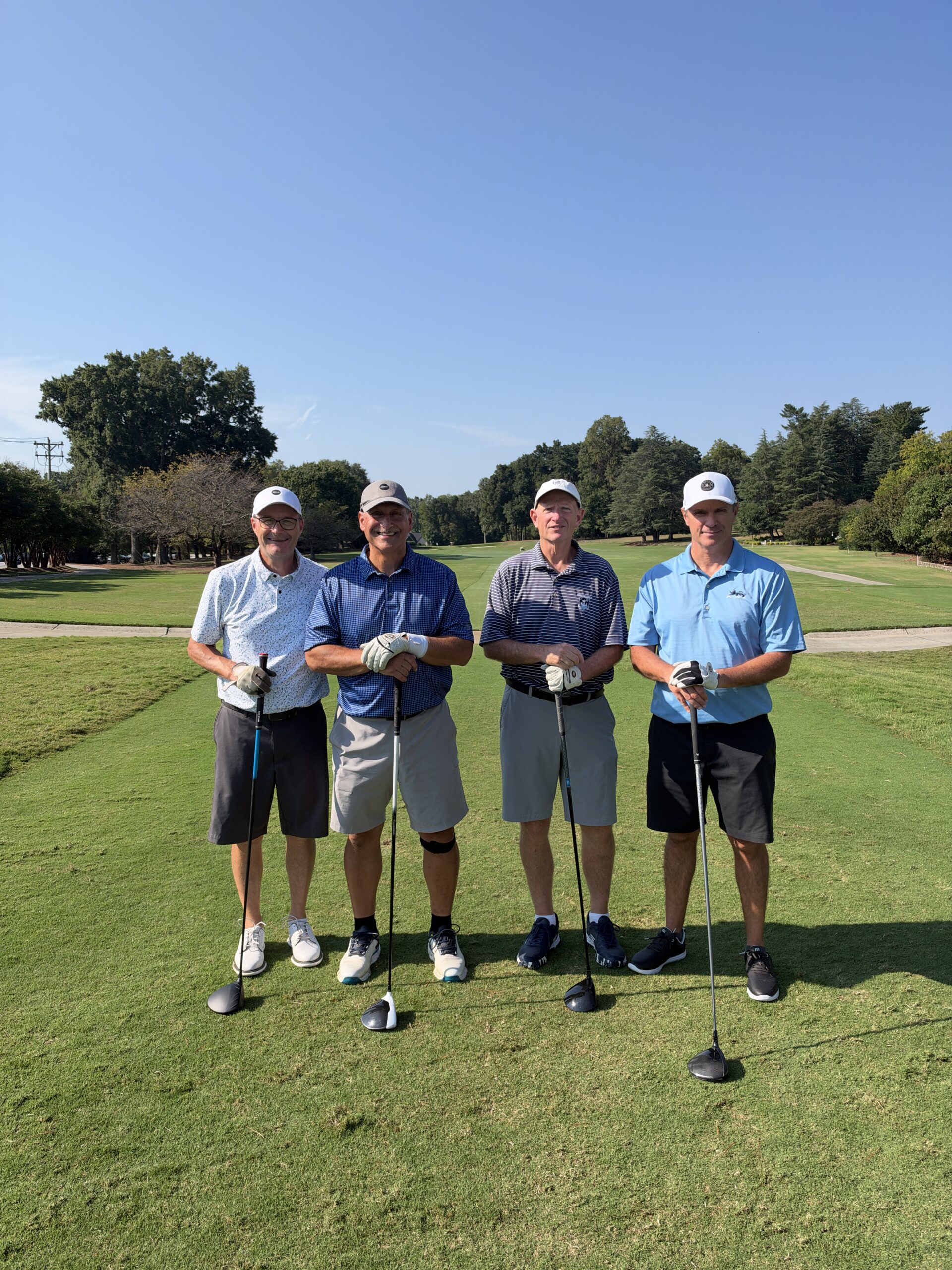 Four men in golf attire stand smiling on a sunny fairway, each leaning on a golf club.