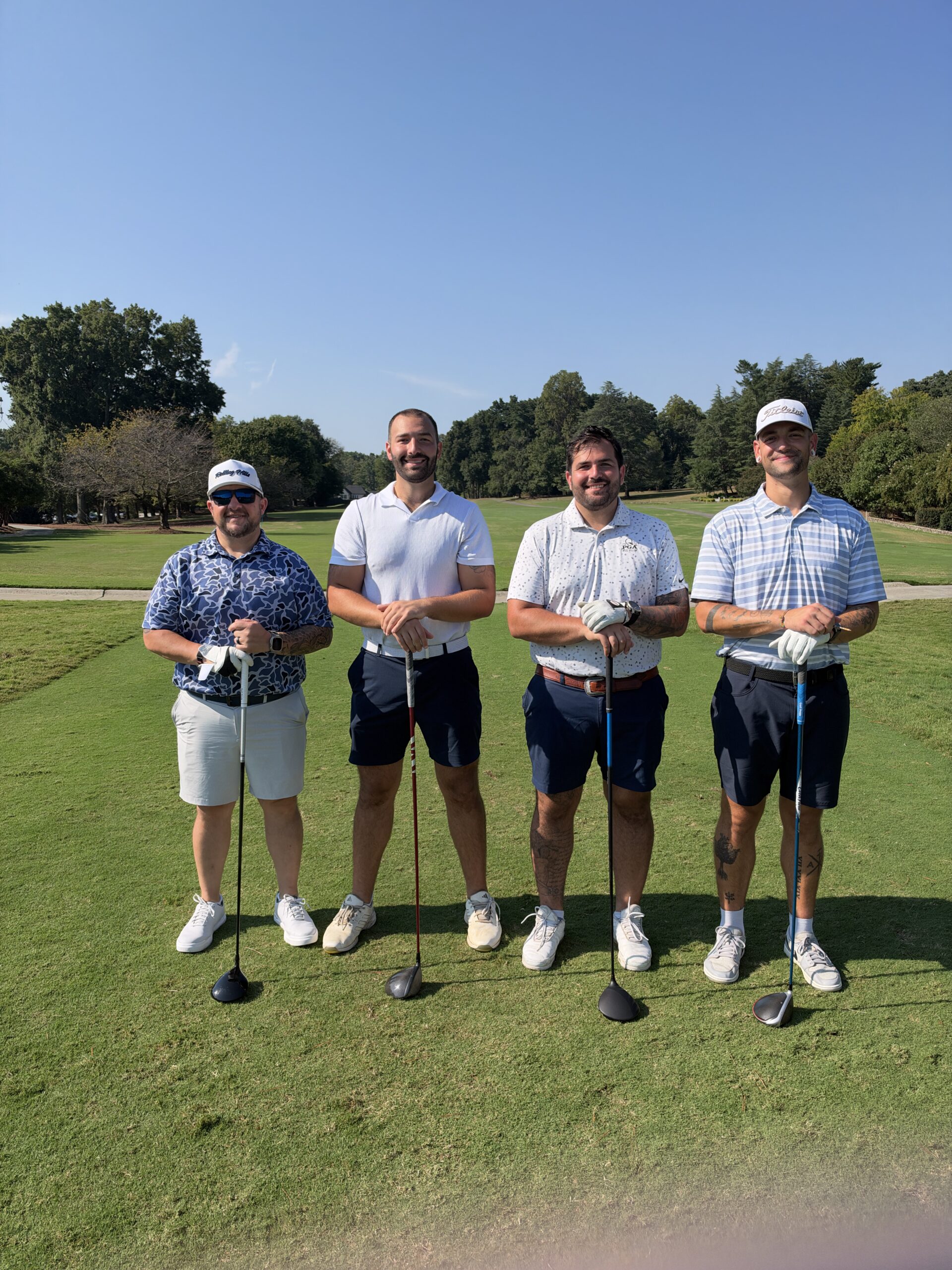 Four men in golf attire pose on a sunny fairway, each leaning on a golf club, with trees behind them.