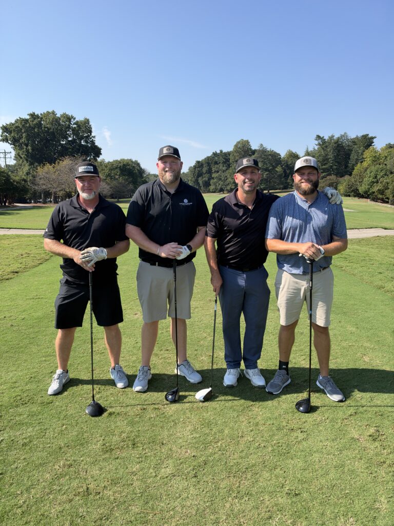 Four men in golf attire pose with clubs on the course at the BCH Golf Classic at Sedgefield Country Club.