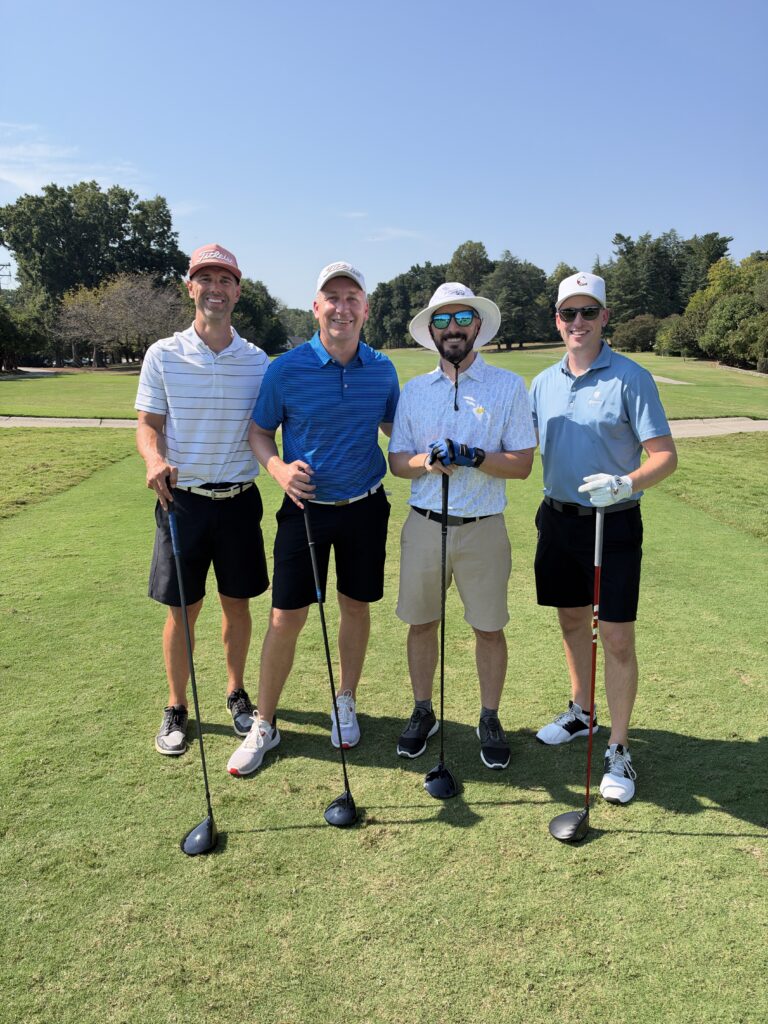 Four smiling men pose with golf clubs on a sunny fairway at the BCH Golf Classic at Sedgefield Country Club.