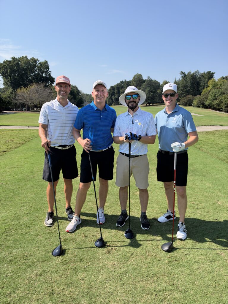 Four men in golf attire pose with clubs on the course at the BCH Golf Classic at Sedgefield Country Club.