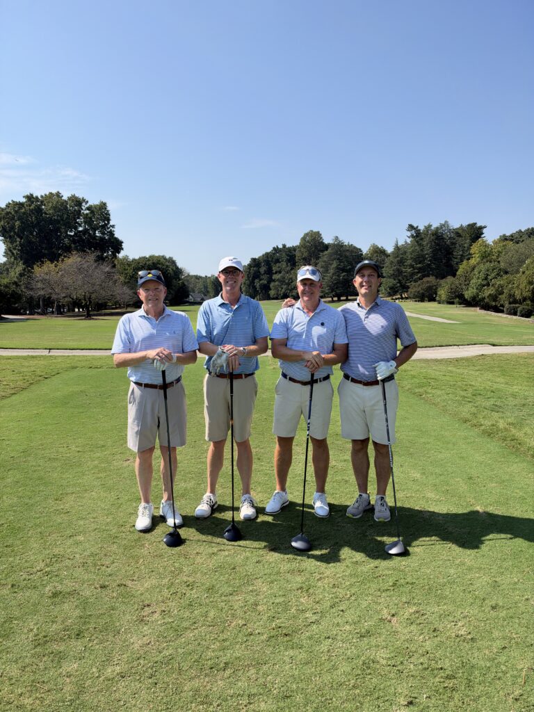 Four men in polos and khaki shorts pose with golf clubs on a fairway at the BCH Golf Classic at Sedgefield Country Club.