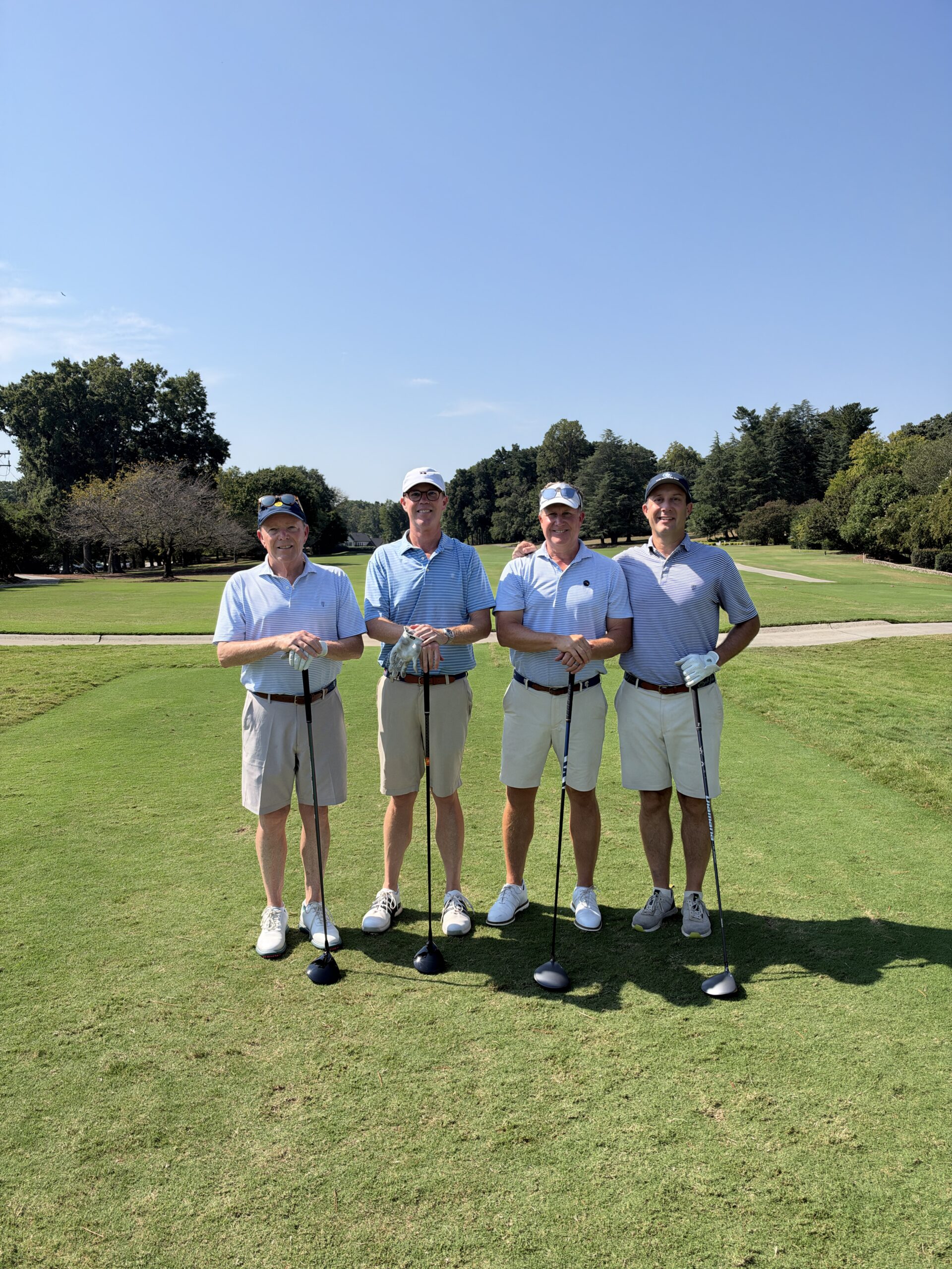 Four men in golf shirts and shorts pose with drivers on a sunny fairway at the BCH Golf Classic at Sedgefield Country Club.
