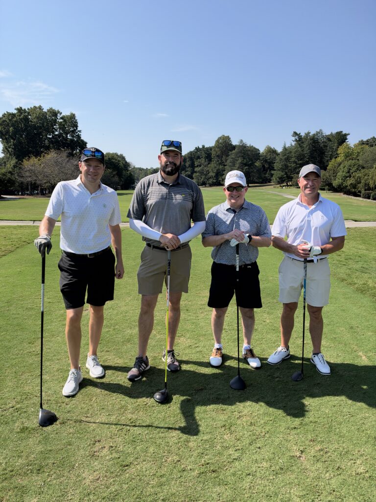 Four men in golf attire pose with clubs on a fairway at the BCH Golf Classic at Sedgefield Country Club.