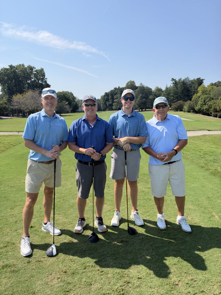 Four men in golf attire stand smiling on the course holding clubs at the BCH Golf Classic at Sedgefield Country Club.