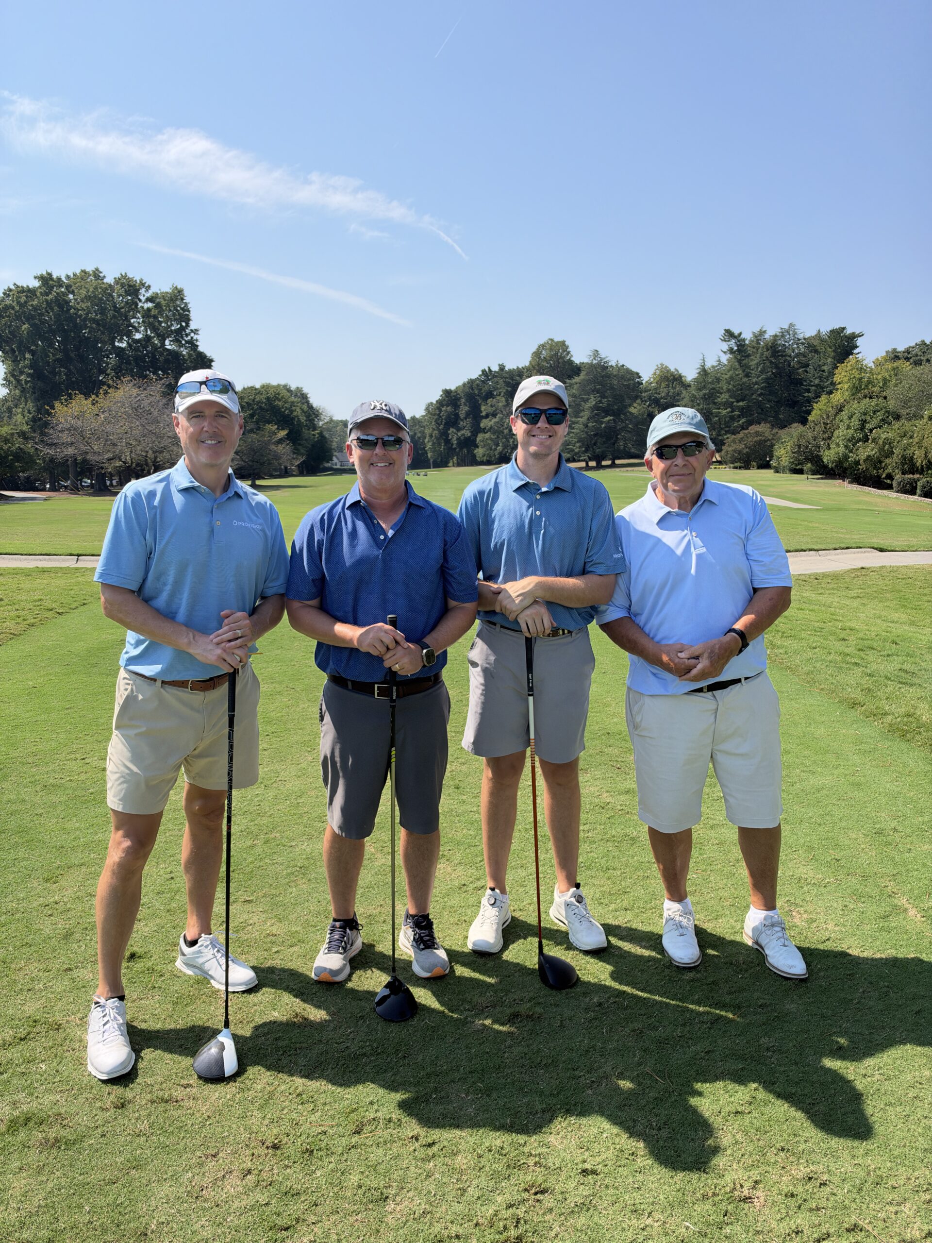 Four men in golf attire stand smiling on the course holding clubs at the BCH Golf Classic at Sedgefield Country Club.