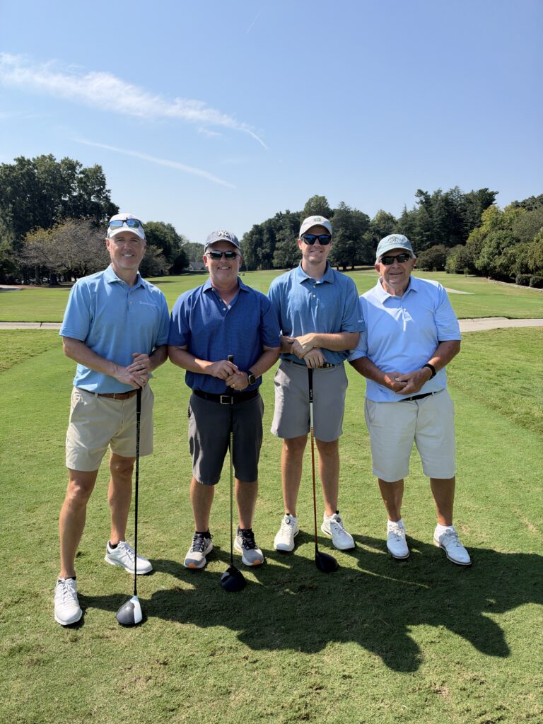 Four men in blue polos and shorts pose with golf clubs on the course at the BCH Golf Classic at Sedgefield Country Club.