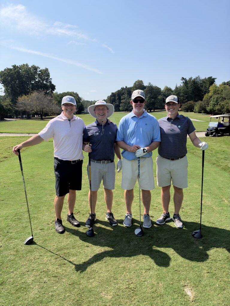 Four men pose smiling with golf clubs on a sunny fairway at the BCH Golf Classic at Sedgefield Country Club.