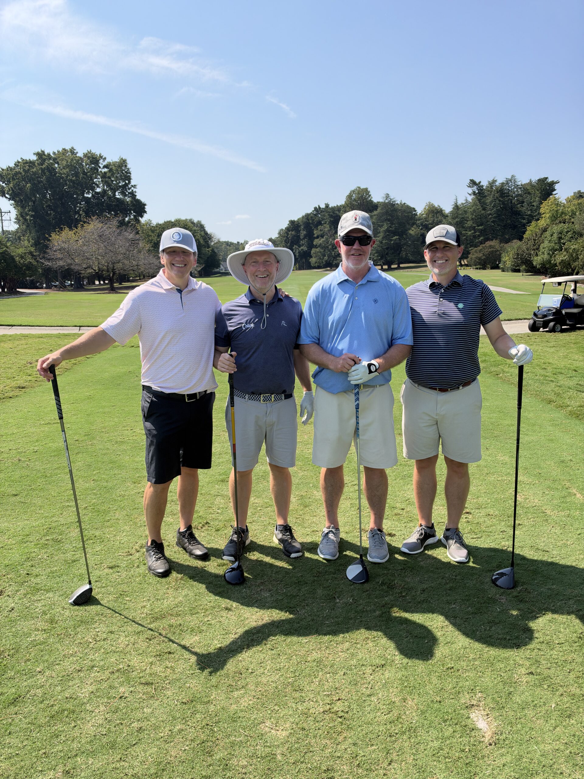 Four men in golf attire pose with drivers on a sunny fairway at the BCH Golf Classic at Sedgefield Country Club.