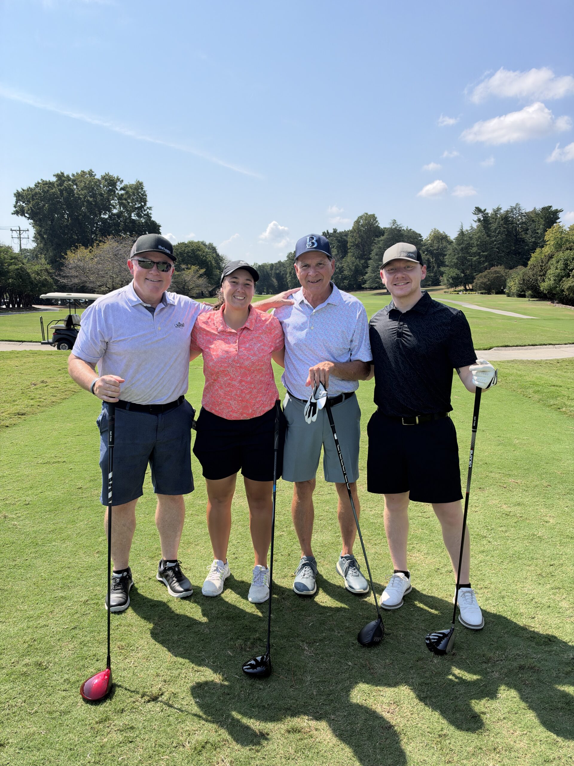 Four golfers pose arm in arm on a sunny fairway holding drivers at the BCH Golf Classic at Sedgefield Country Club.