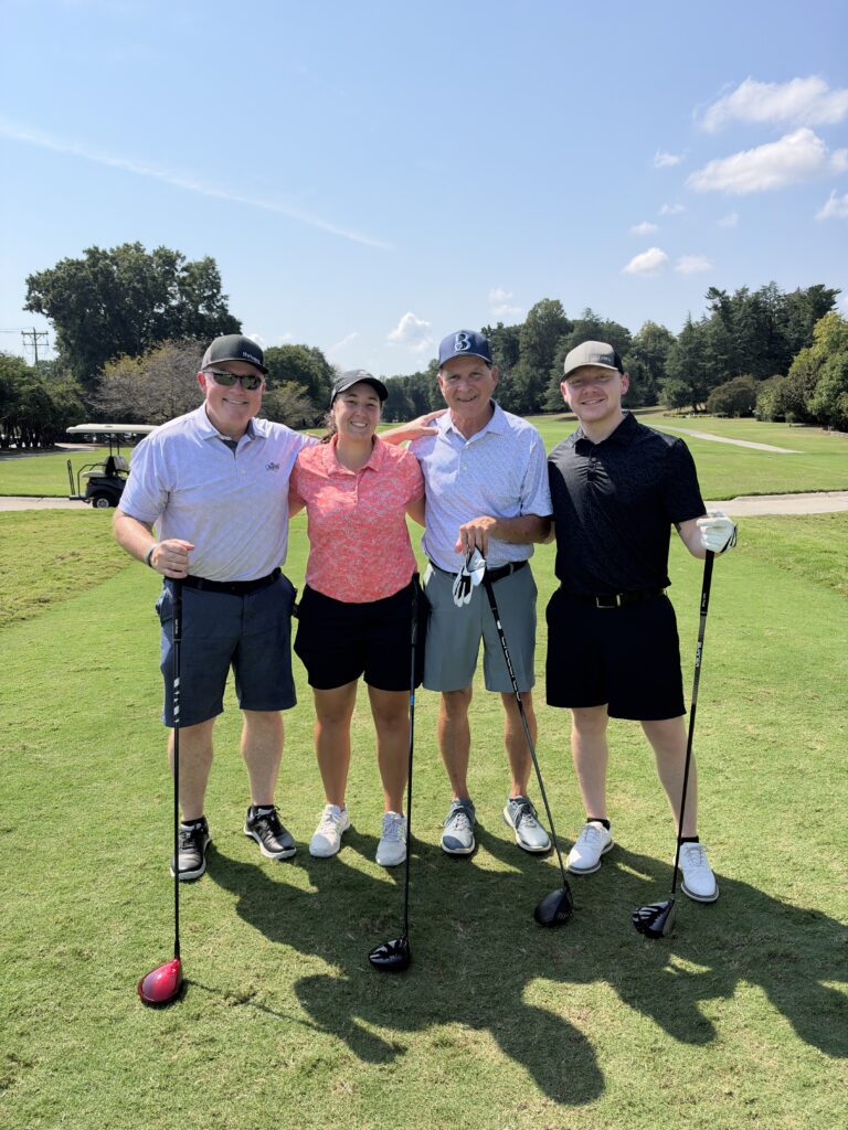 Four golfers pose arm in arm on a sunny fairway holding clubs at the BCH Golf Classic at Sedgefield Country Club.