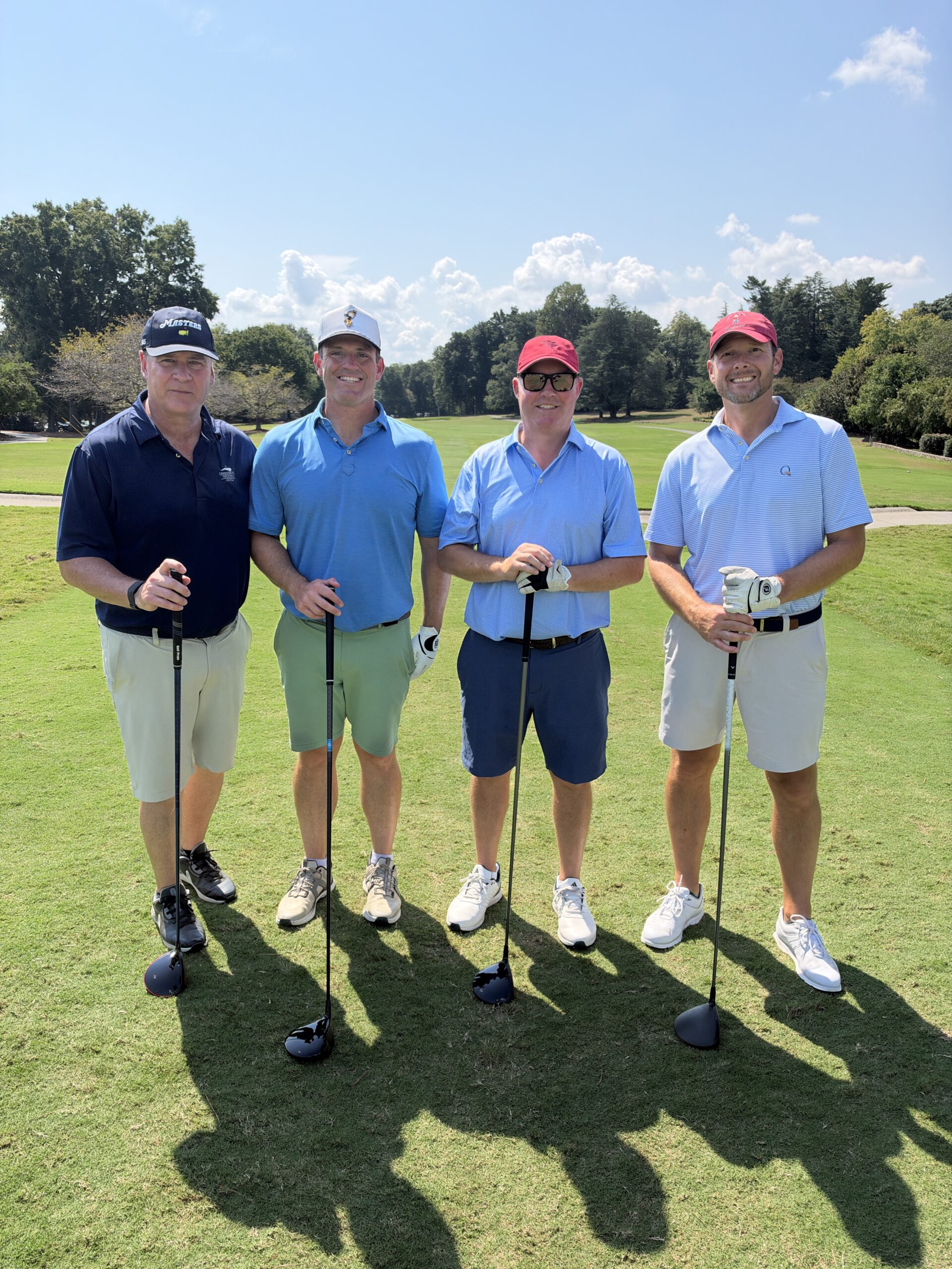 Four men in golf attire pose with drivers on the course at the BCH Golf Classic at Sedgefield Country Club.