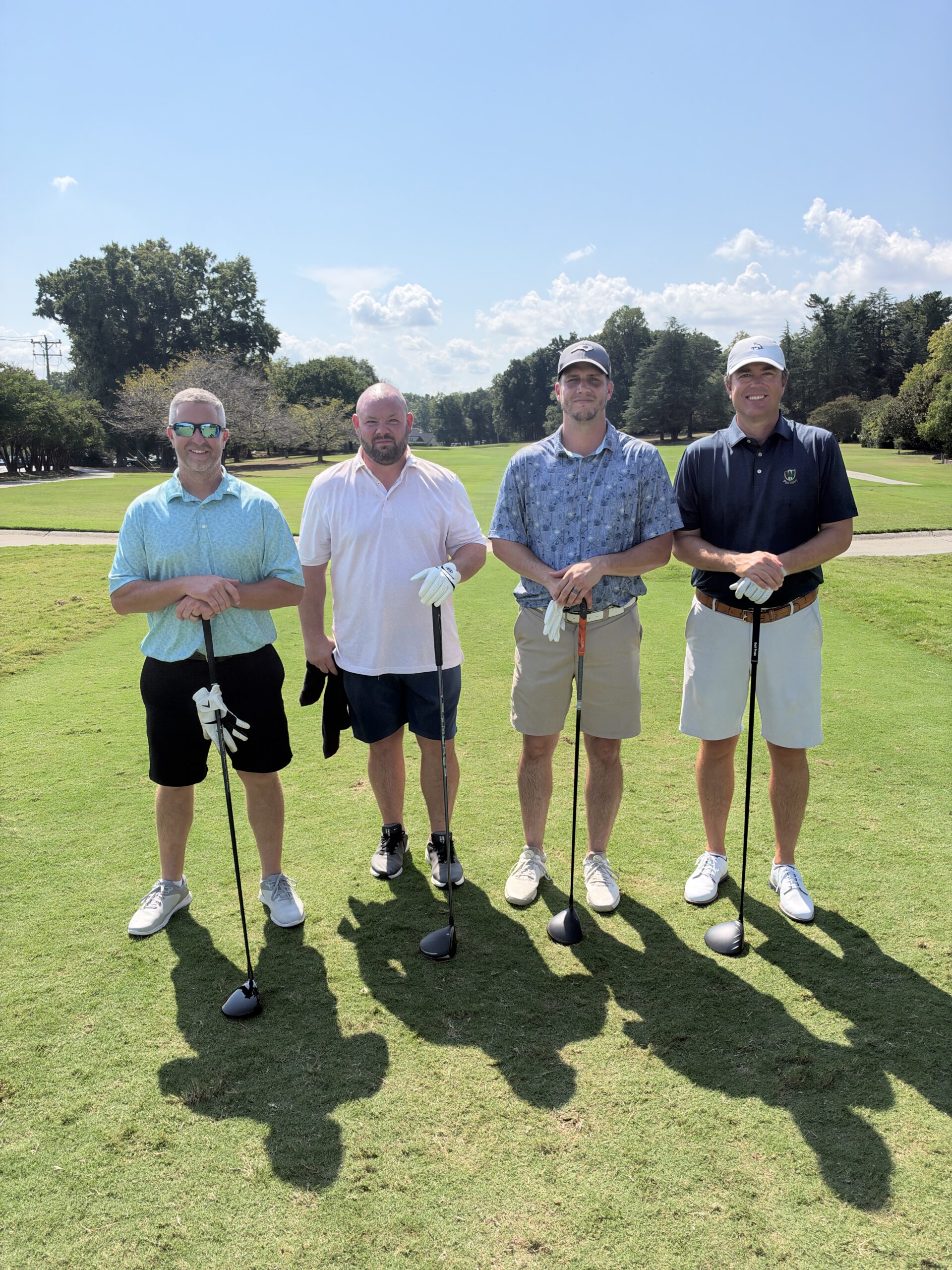 Four men pose with golf clubs on the course at the BCH Golf Classic fundraising tournament at Sedgefield Country Club.