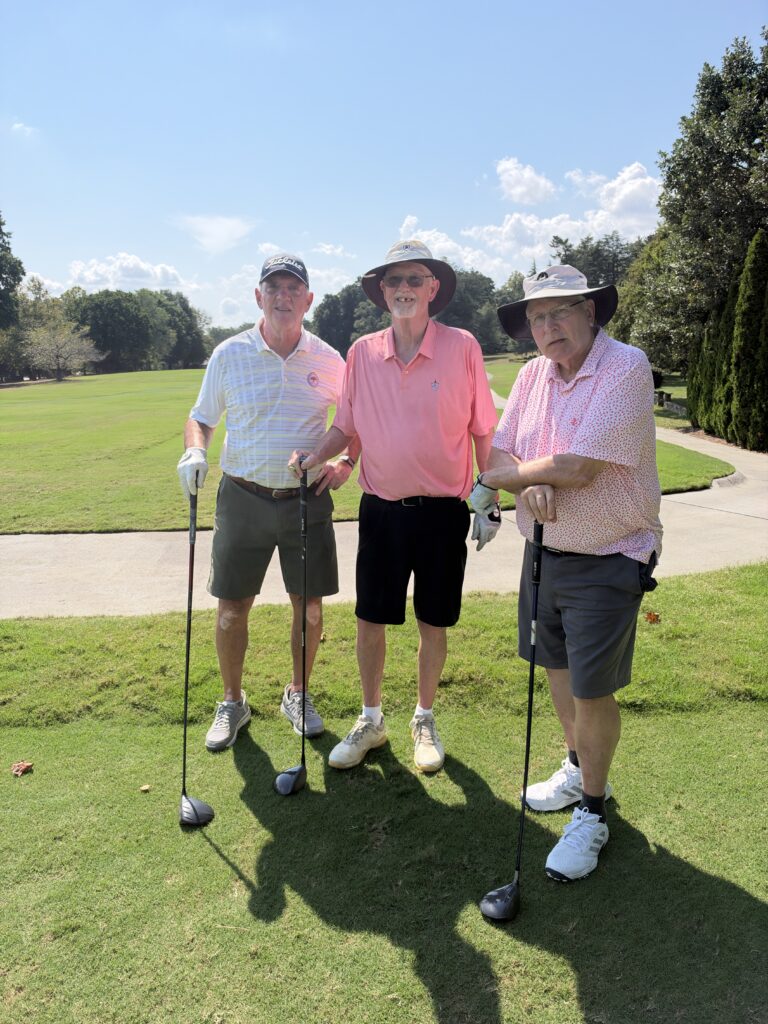 Three men in golf attire stand smiling on a course holding clubs at the BCH Golf Classic at Sedgefield Country Club.