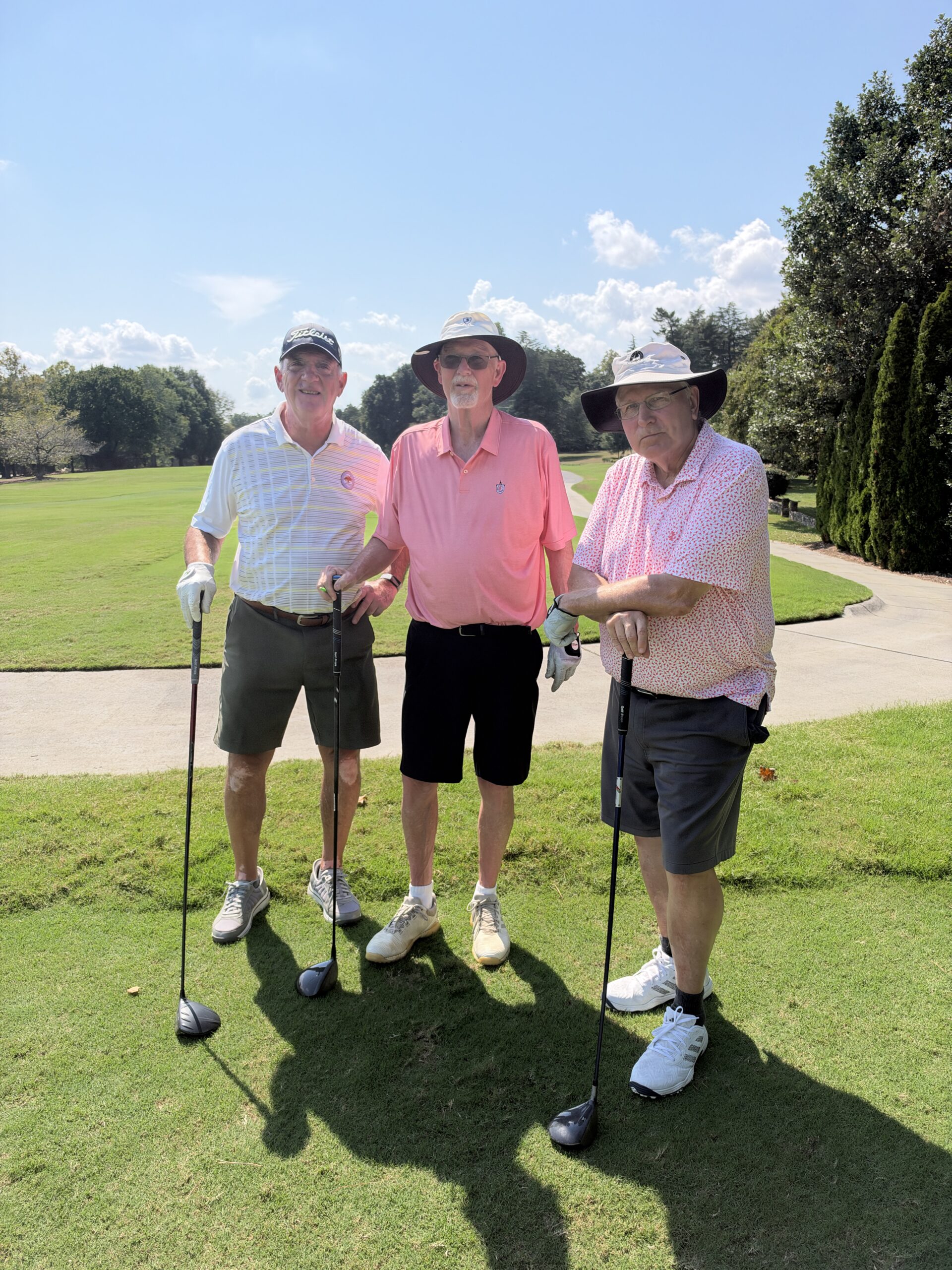 Three men in golf attire pose with their clubs on a sunny course at the BCH Golf Classic at Sedgefield Country Club.