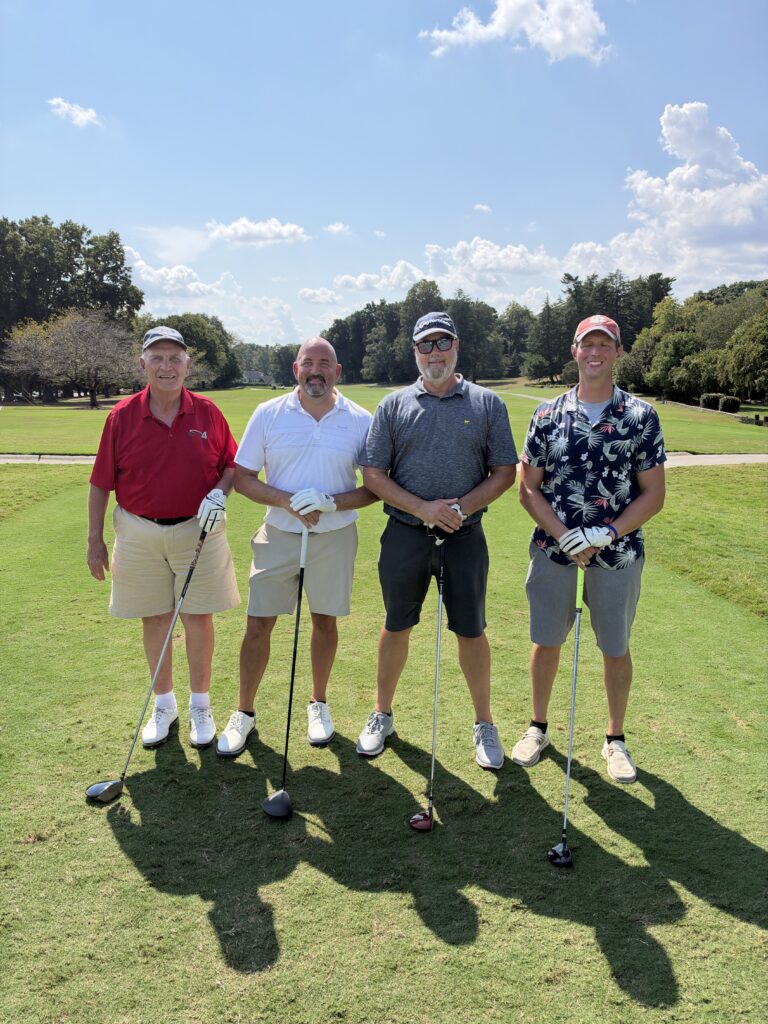 Four men pose with golf clubs on a sunny fairway at the BCH Golf Classic fundraising tournament at Sedgefield Country Club.