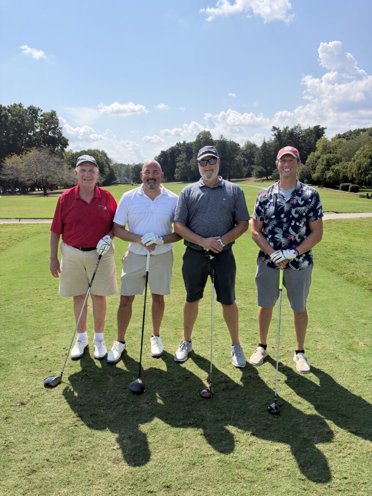 Four men pose with golf clubs on a sunny fairway at the BCH Golf Classic fundraising tournament at Sedgefield Country Club.