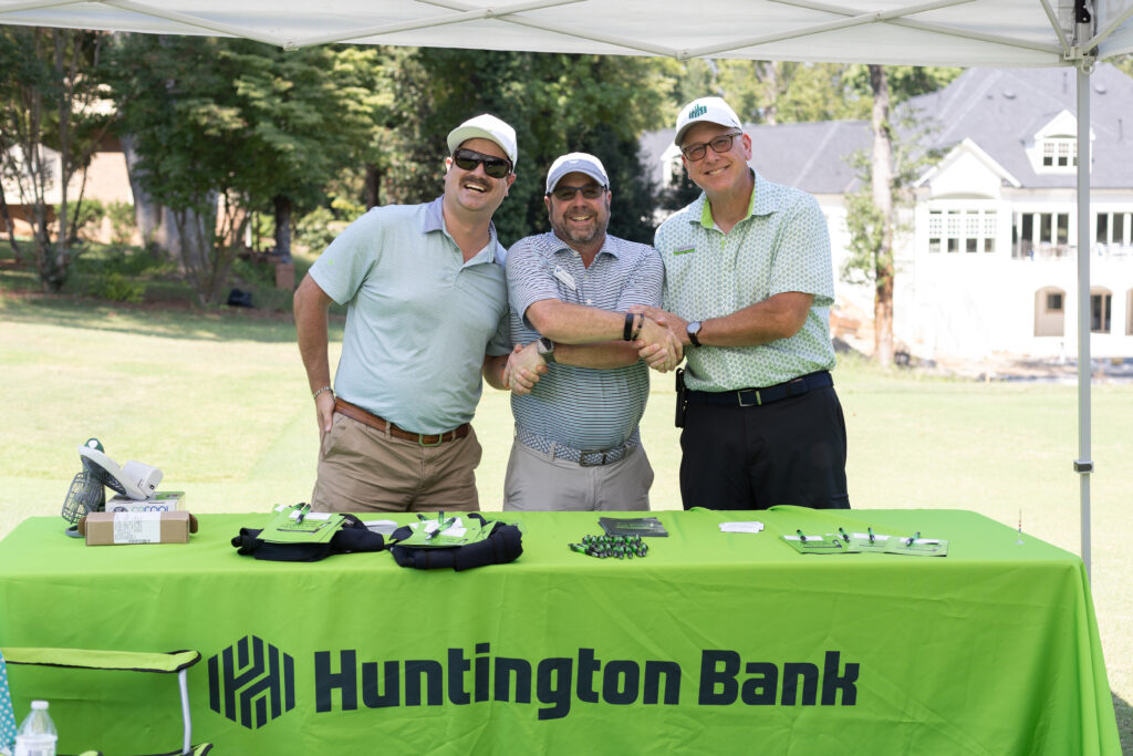 Three men in golf attire clasp hands at a Huntington Bank table during the BCH Golf Classic at Sedgefield Country Club.