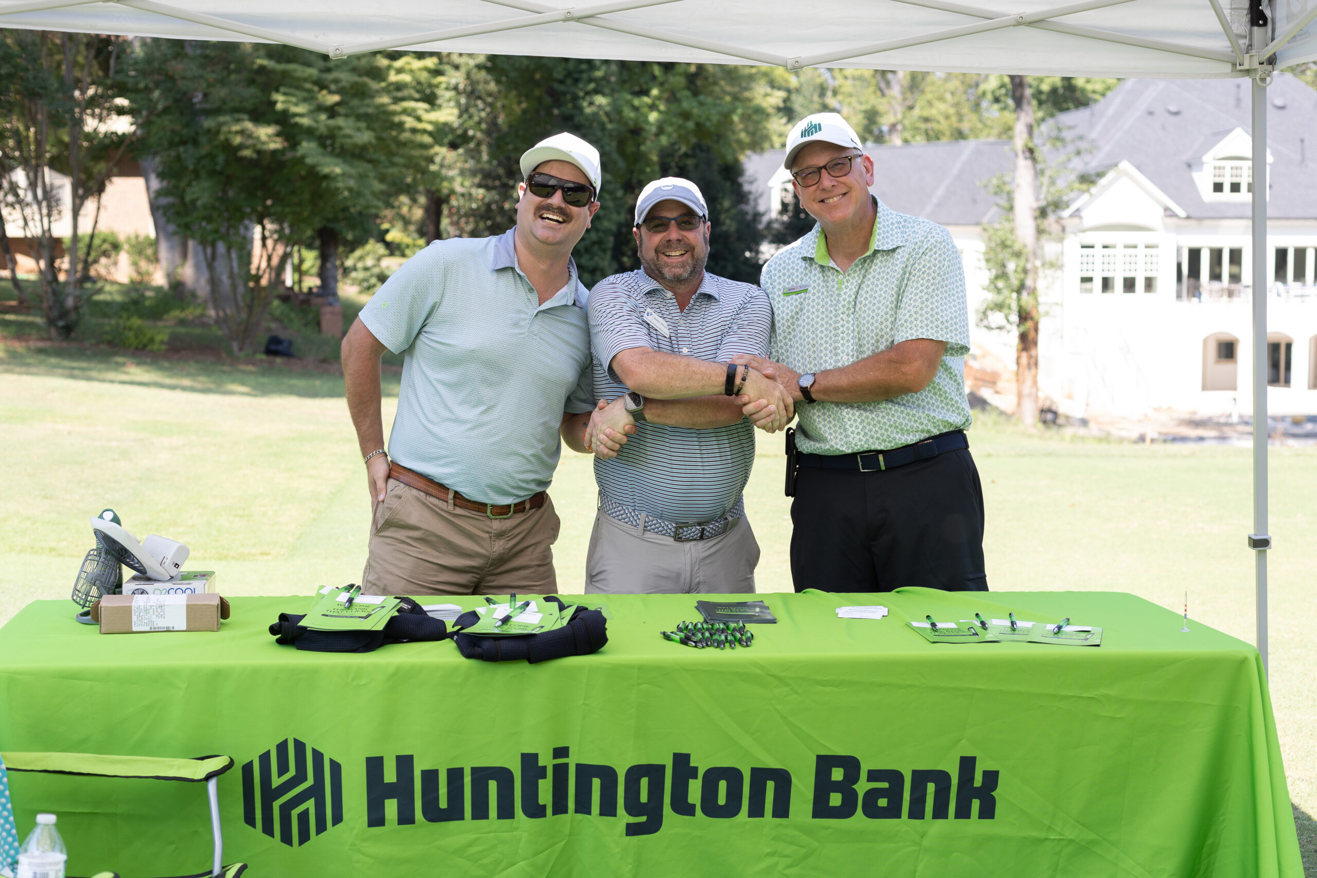 Three men in golf attire clasp hands at a Huntington Bank table during the BCH Golf Classic at Sedgefield Country Club.