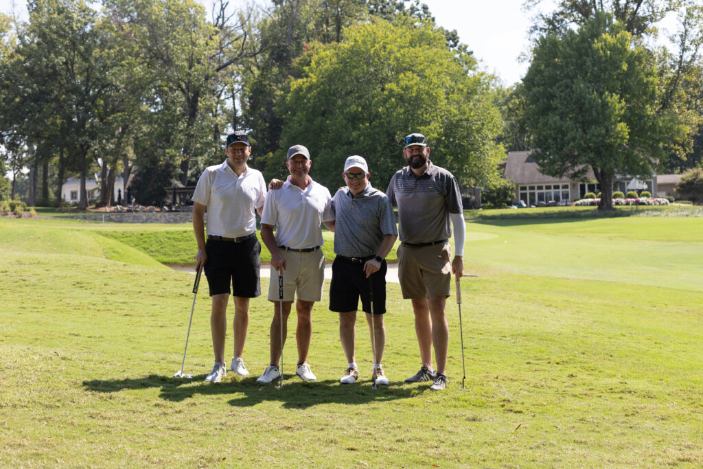 Four men in golf attire pose together holding clubs on a sunny fairway during the BCH Golf Classic at Sedgefield Country Club.