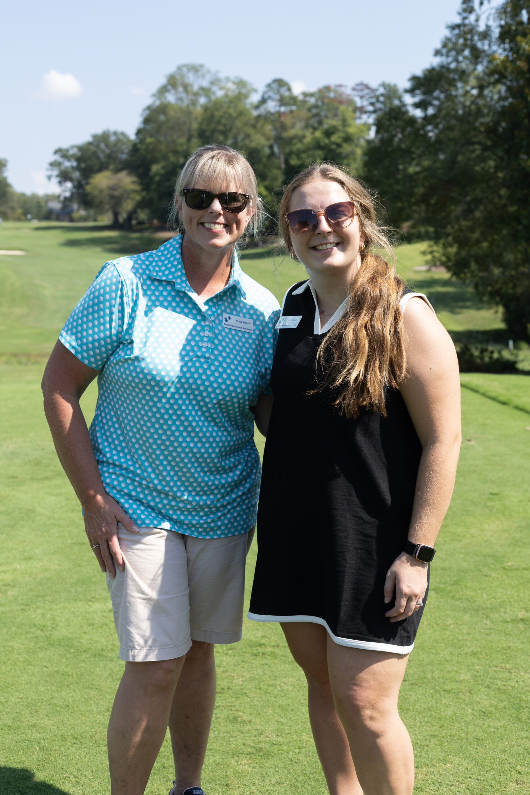 Two smiling women in sunglasses wearing name tags pose on a golf course at the BCH Golf Classic at Sedgefield Country Club.