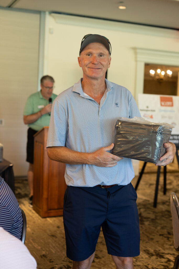 Smiling man in a blue polo and cap holds a wrapped gift at the BCH Golf Classic while a speaker stands at a podium behind him.