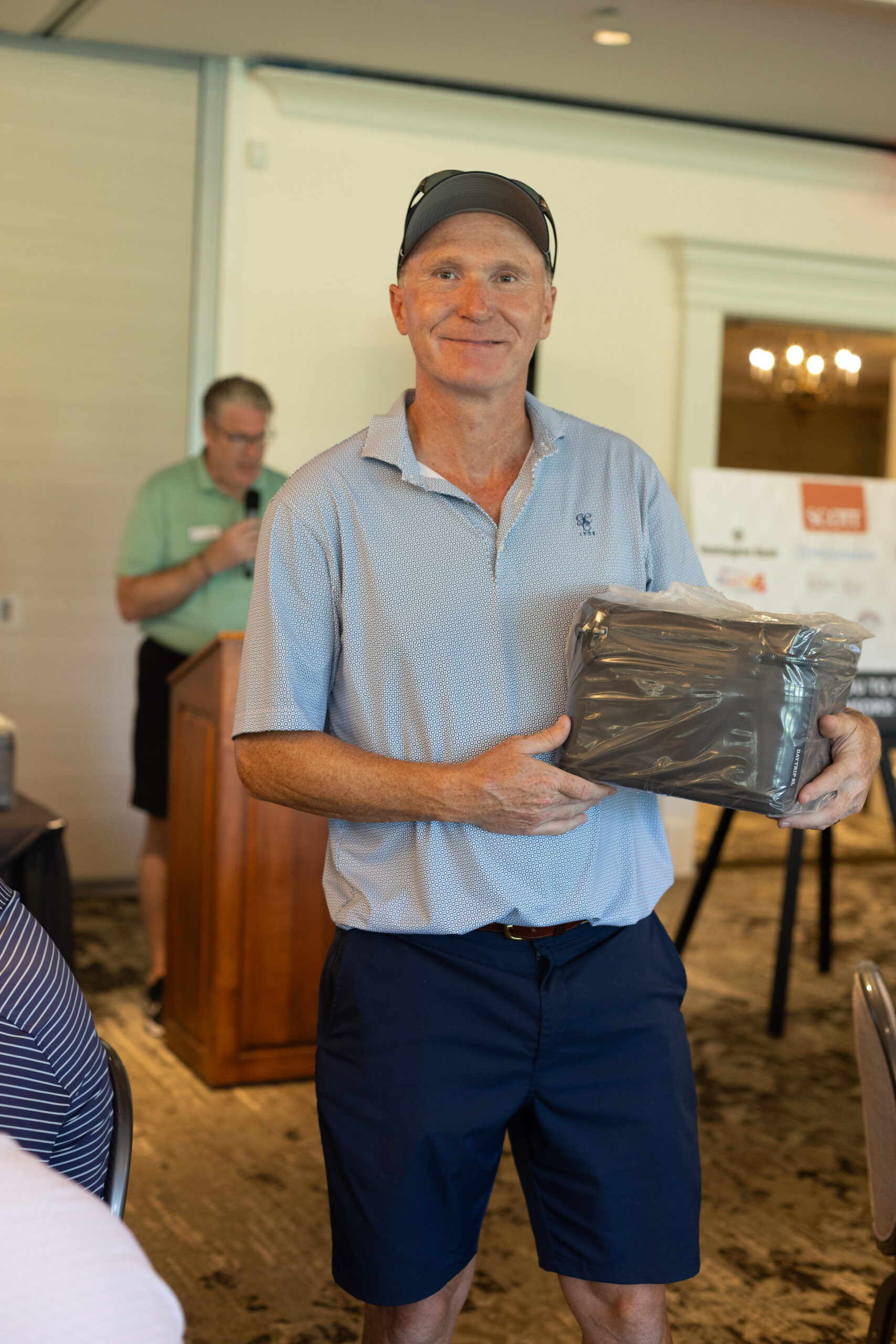 Smiling man in a blue polo and cap holds a wrapped gift at the BCH Golf Classic while a speaker stands at a podium behind him.