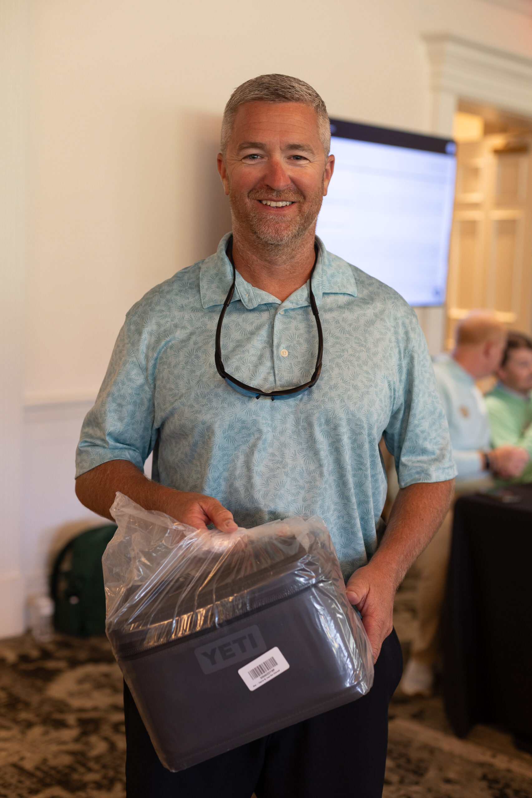 Smiling man in a light blue polo holds a YETI cooler in plastic wrap at the BCH Golf Classic at Sedgefield Country Club.