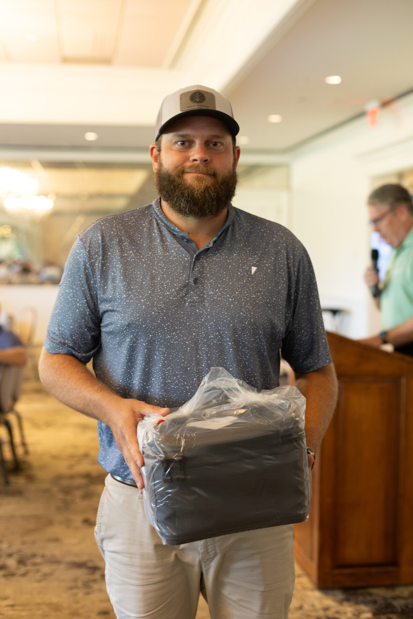 Bearded man in a cap and blue polo holds a prize in plastic wrap at the BCH Golf Classic at Sedgefield Country Club.
