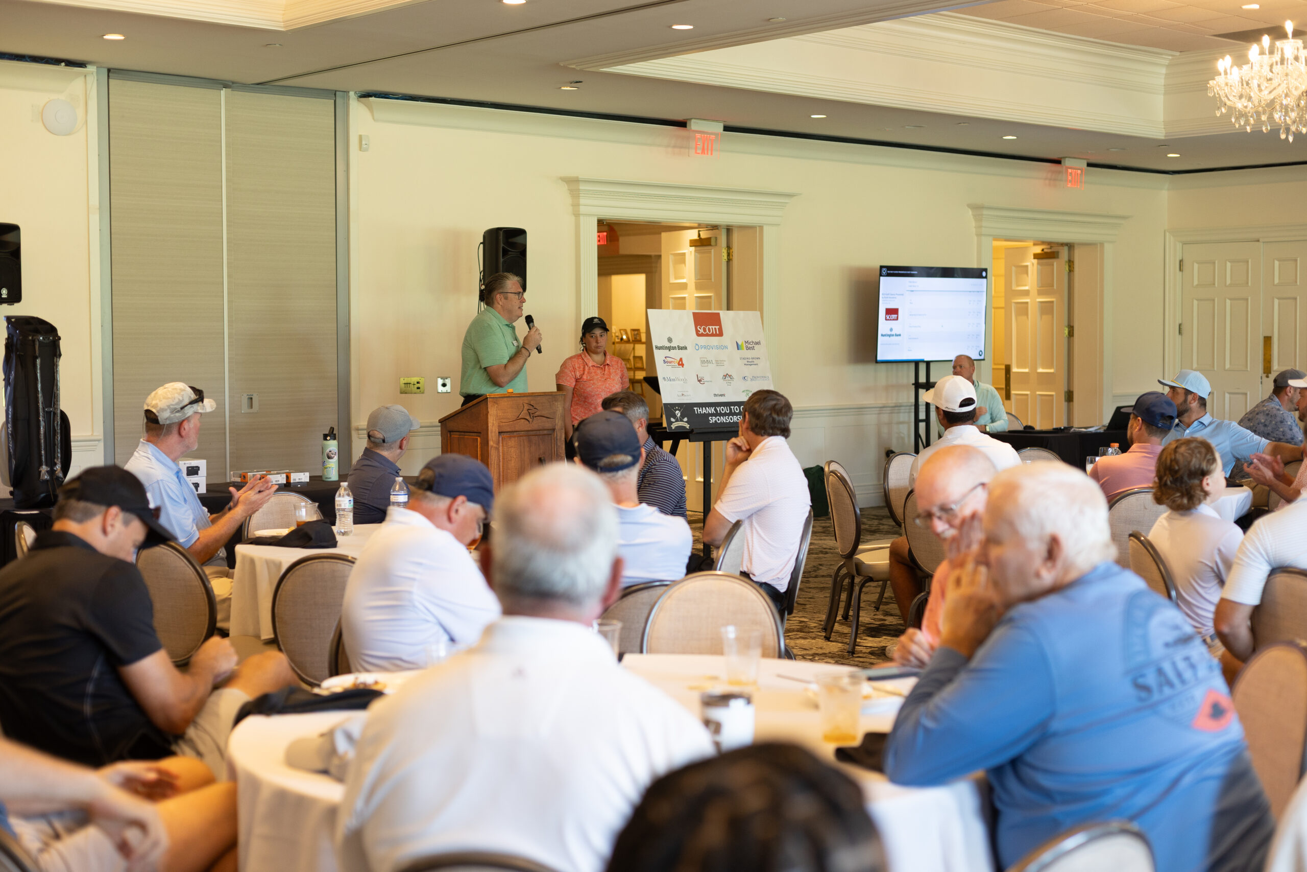 Speaker at a podium addresses golfers seated at tables during the BCH Golf Classic, with a sponsor thank-you sign nearby.