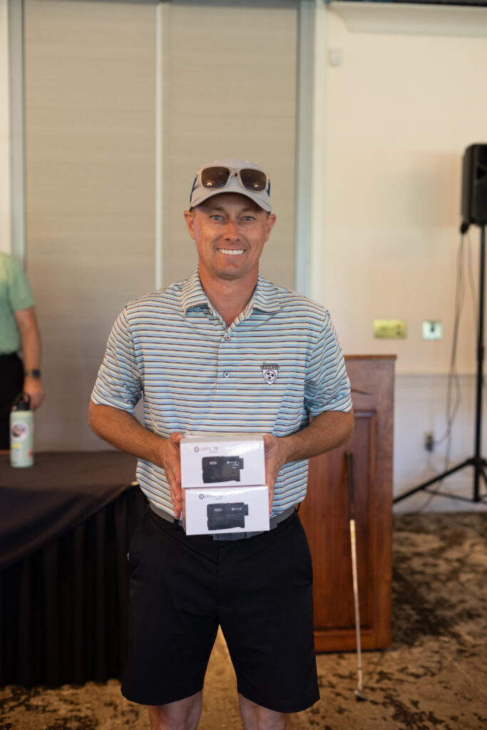 Smiling man in a striped polo and cap holds two boxed golf rangefinders at the BCH Golf Classic at Sedgefield Country Club.