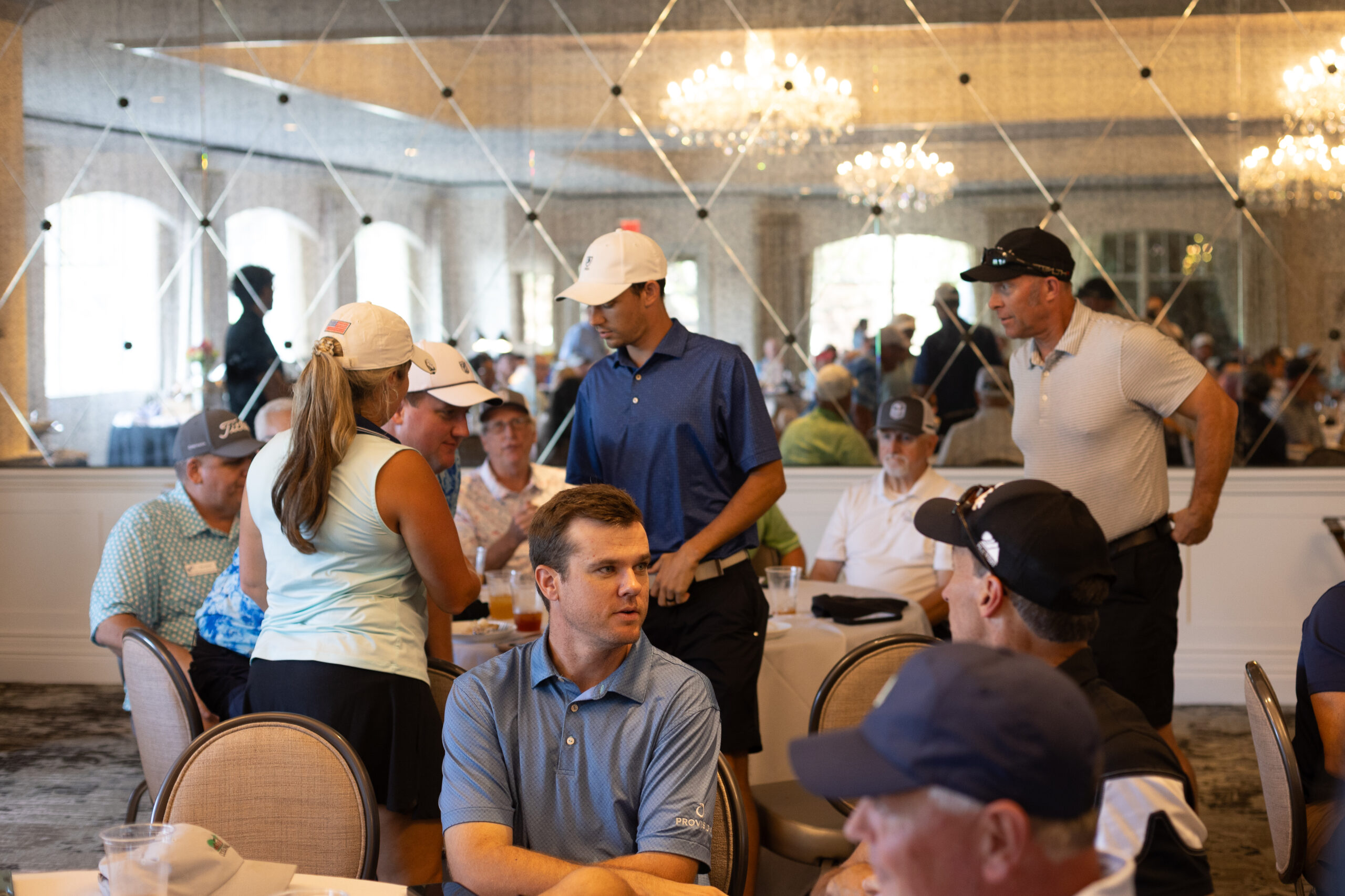 Golfers in polo shirts and caps talk at round tables in a chandeliered ballroom during the BCH Golf Classic at Sedgefield.