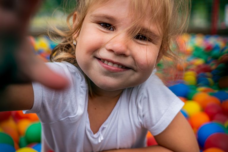 A happy child smiling and reaching towards the camera in a vibrant ball pit outdoors.
