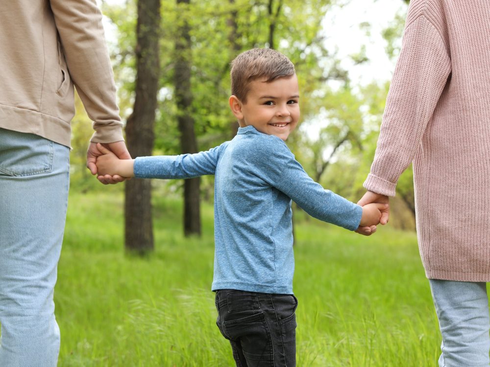 A smiling boy walks hand in hand with two adults through a park